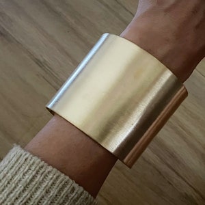 Puede incluir: Un brazalete ancho de metal dorado. El brazalete es liso y tiene una forma ligeramente redondeada.