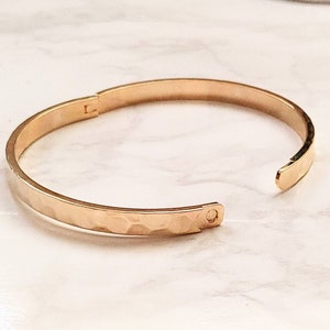 May include: A gold-toned hammered metal bangle bracelet. The bracelet is open and has a hinged clasp.