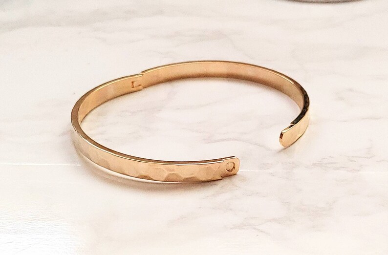 May include: A gold-toned hammered metal bangle bracelet. The bracelet is open and has a hinged clasp.