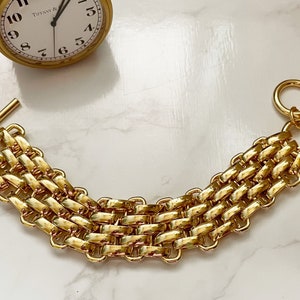 Op de afbeelding: Een gouden kettingarmband met een geweven ontwerp en een sluiting. De armband is gemaakt van dikke, stevige schakels.