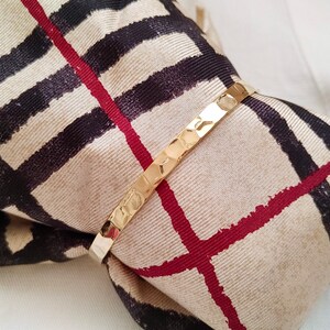 May include: A gold bangle bracelet with a textured surface. The bracelet is wrapped around a beige and black patterned fabric.