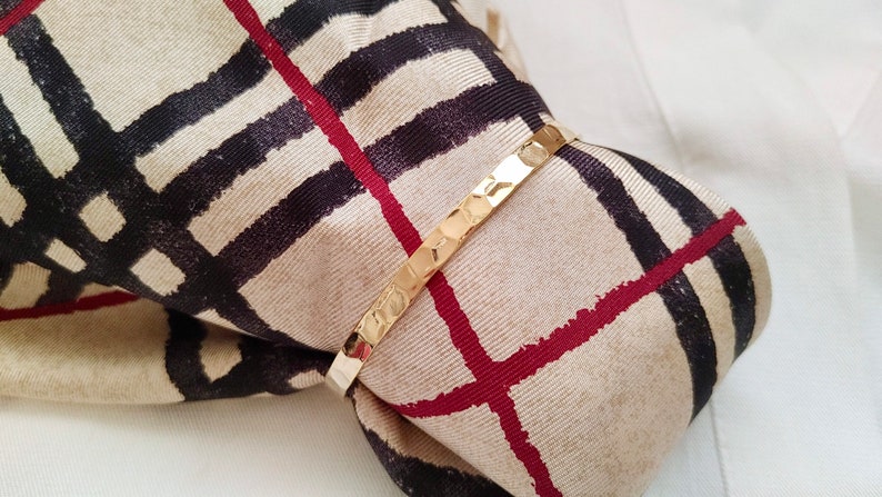 May include: A gold bangle bracelet with a textured surface. The bracelet is wrapped around a beige and black patterned fabric.