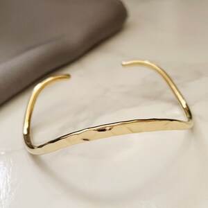 May include: A gold-toned hammered metal bangle bracelet. The bracelet is open and has a simple, minimalist design.