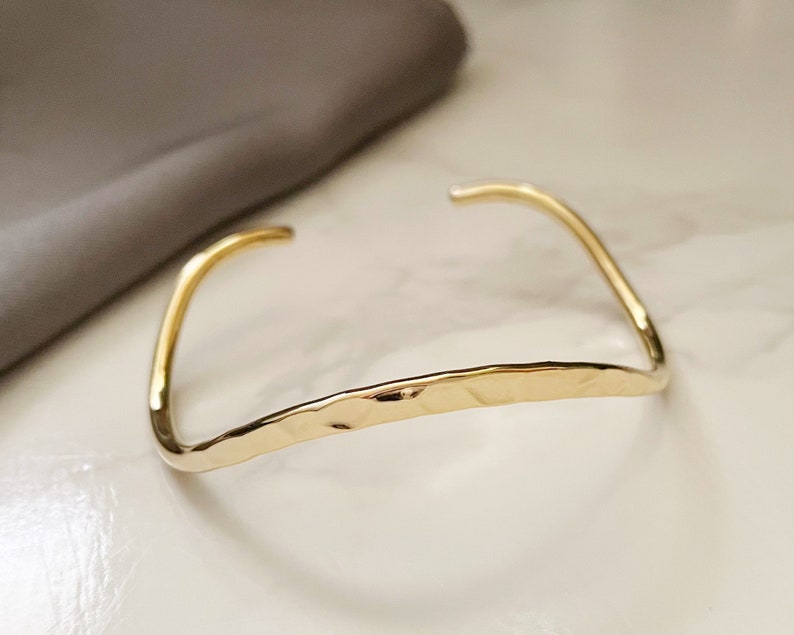 May include: A gold-toned hammered metal bangle bracelet. The bracelet is open and has a simple, minimalist design.