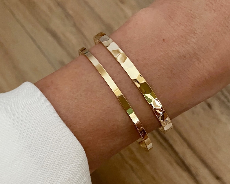 May include: Two gold bracelets on a wrist. One bracelet is a simple, thin band, while the other is a thicker band with a hammered texture.