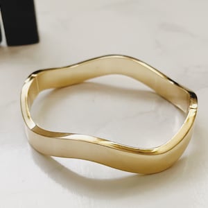 Può includere: Un braccialetto rigido ondulato color oro. Il braccialetto è realizzato in metallo liscio e lucido e ha un design semplice ed elegante.