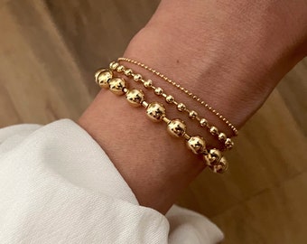 Può includere: Un set di tre braccialetti d'oro, uno con una catena delicata e due con perline d'oro graduate.