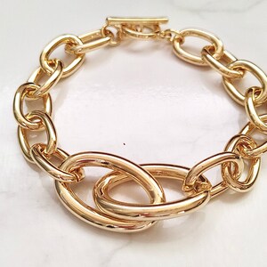 Op de afbeelding: Een gouden kettingarmband met grote, ovale schakels. De armband is gemaakt van dik, robuust metaal en heeft een eenvoudig, modern ontwerp.