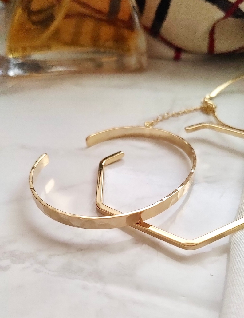 May include: Two gold bracelets, one with a hammered finish and the other a geometric hexagon shape. The bracelets are linked together with a delicate chain.