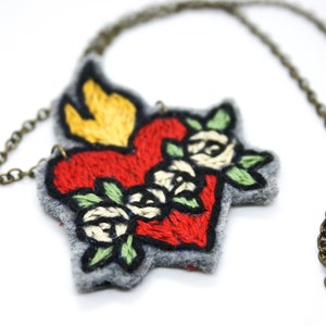 May include: A handmade necklace featuring a felt pendant in the shape of a heart with a flame and roses. The heart is red with yellow flames and white roses, outlined in black, and attached to a bronze chain.