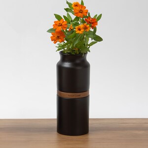 May include: A black ceramic vase with a brown leather band that says "Forever Loved" holds a bouquet of orange flowers.