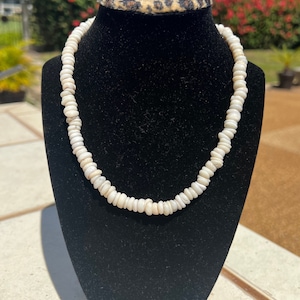 May include: A white shell necklace on a black velvet display stand. The necklace is made of small, round shells that are strung together.