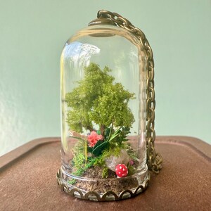 May include: A miniature terrarium in a glass cloche with a brass-coloured chain. It contains a small tree, pink flowers, moss, and a red and white mushroom. The cloche is displayed on a brown surface.