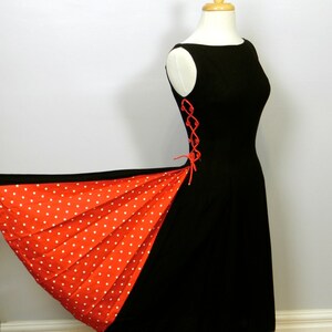May include: A sleeveless black dress featuring a side panel with red and white polka dots. The side of the bodice includes a red lace-up detail with small red buttons. The skirt flares out to reveal the pleated red and white polka dot fabric.