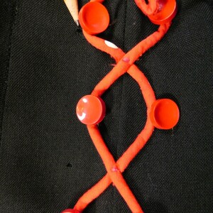 May include: A black fabric garment featuring a decorative crisscross pattern made of orange fabric tubing. The design is accented with six round, bright orange buttons attached at the intersections of the tubing. A yellow pencil is visible.