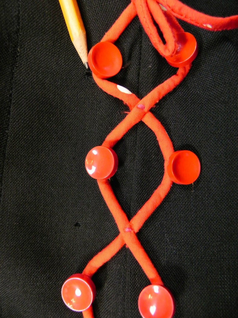 May include: A black fabric garment featuring a decorative crisscross pattern made of orange fabric tubing. The design is accented with six round, bright orange buttons attached at the intersections of the tubing. A yellow pencil is visible.