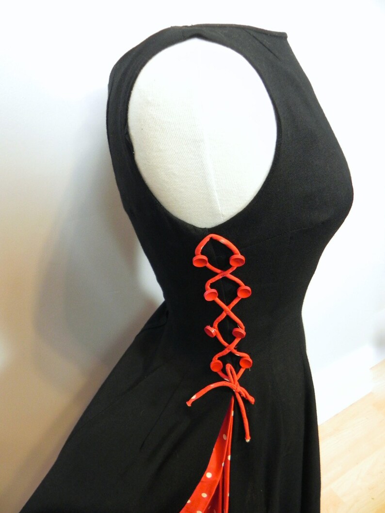 May include: A sleeveless black dress featuring a side lace-up detail with bright orange buttons and matching orange cord lacing. The side opening reveals a red fabric panel with white polka dots underneath the lacing and a tied cord at the base.