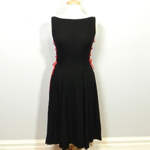 May include: A sleeveless black dress featuring a boat neckline and a fitted bodice that flares into a pleated skirt. The sides of the dress are accented with vertical rows of bright red buttons and decorative red lacing details along the seams.
