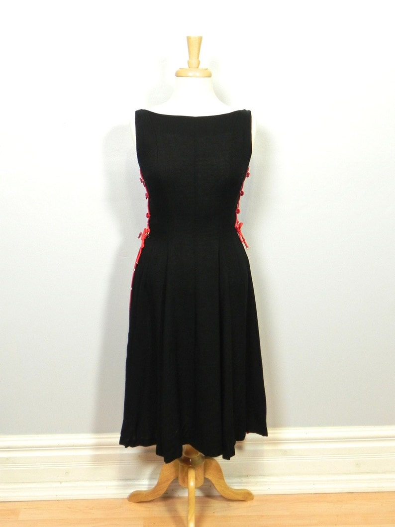 May include: A sleeveless black dress featuring a boat neckline and a fitted bodice that flares into a pleated skirt. The sides of the dress are accented with vertical rows of bright red buttons and decorative red lacing details along the seams.