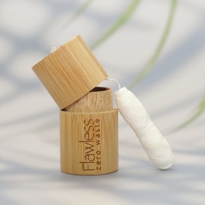 May include: A bamboo floss container with the text "Flawless zero waste" printed on the side. The container is open, revealing a spool of white floss. The container and lid are made of light brown bamboo.