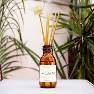 Könnte beinhalten: Eine braune Glasflasche mit einem weißen Etikett, auf dem "Flawless* HAPPINESS reed diffuser" steht, und einer weißen Blume oben drauf. Die Flasche ist mit einer klaren Flüssigkeit gefüllt und hat einen Satz Holzstäbchen, die hineingesteckt sind.