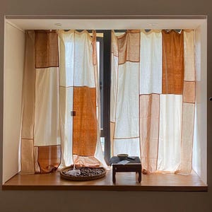 Puede incluir: Una ventana con cortinas de patchwork blanco y marrón. Las cortinas están hechas de una tela ligera y tienen un estilo rústico, de granja. La ventana está enmarcada por un marco blanco y está ubicada en una pared con un piso de madera marrón claro.