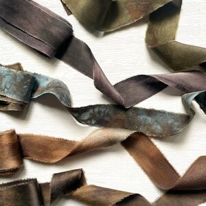 May include: Close-up of several spools of dyed velvet ribbon in various shades of brown, blue, and green. The ribbons have a soft, textured appearance and are arranged in a flowing, organic pattern. The edges of the ribbon are frayed.