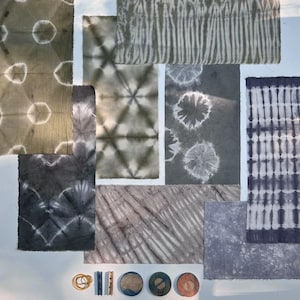 Op de afbeelding: Een assortiment stoffen stalen met verschillende tie-dye patronen in grijstinten, blauw en olijfgroen. De ontwerpen omvatten cirkels, strepen en geometrische vormen. Er zijn ook elastiekjes, wasknijpers en houten schijven.