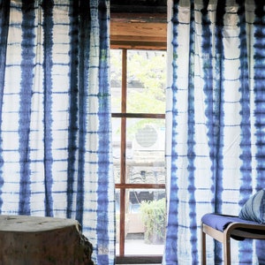 Puede incluir: Cortinas de tie-dye azul y blanco con una ventana detrás. Las cortinas tienen un patrón de rayas con un fondo blanco y rayas azules.