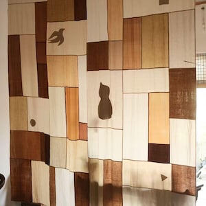 Puede incluir: Un gran tapiz rectangular de tela con un diseño de patchwork compuesto por varios paneles rectangulares en tonos crema, beige, tostado y marrón oscuro. El tejido incluye pequeñas siluetas oscuras de un pájaro, un gato, un círculo y un triángulo.