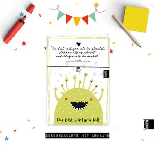 Könnte beinhalten: Ein grünes Monster mit mehreren Augen und einem großen Lächeln auf einer weißen Karte mit dem Text "Du bist mutiger als Du glaubst, starker als es scheint und kluger als Du denkst" und "Du bist einfach toll". Die Karte trägt außerdem den Text "Geschenkkarte mit Armband" am unteren Rand.