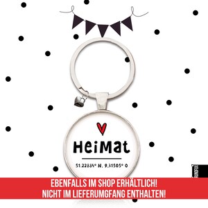 May include: A silver keychain with a round charm that says "Heimat" with a red heart above the word. The charm also includes the coordinates "51.22884&deg; N. 9,31505&deg; 0".