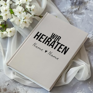 Könnte beinhalten: Weißes Hardcover-Journal mit dem Text "Wir Heiraten" in schwarzer Schrift. Der Text "Name ♥ Name" ist darunter in Kursivschrift geschrieben.