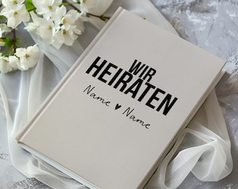 May include: White hardcover journal with the text "Wir Heiraten" in black lettering. The text "Name ♥ Name" is written below in cursive.