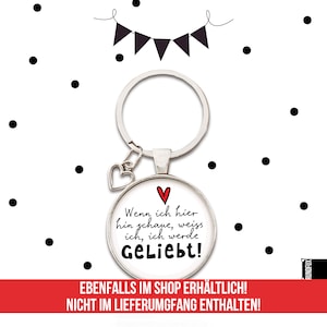 May include: A silver keychain with a round glass charm that says "Wenn ich hier hin schaue, weiss ich, ich werde Geliebt!" in black text with a red heart. The charm is attached to a silver heart charm and a silver key ring.