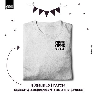 Puede incluir: Una camiseta gris con el texto "YIPPIE YIPPIE YEAH" impreso en la parte delantera. La camiseta es un parche de planchado o una transferencia de planchado que se puede aplicar a la tela.