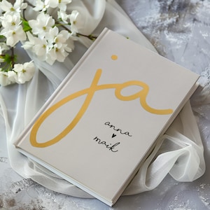 Könnte beinhalten: Ein weißes Hardcover-Journal mit dem Wort "ja" in goldener Schrift. Die Namen "anna" und "maik" stehen unter dem Wort "ja" in schwarzer Schrift mit einem kleinen Herzsymbol zwischen den Namen.