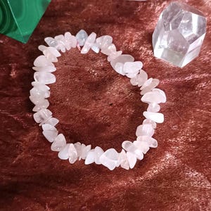 Könnte beinhalten: Ein elastisches Armband aus zahlreichen kleinen, unregelmäßigen und getrommelten hellrosa Rosenquarz-Splittern. Die Steine sind durchscheinend, variieren leicht in Form und Größe und sind zu einem kreisförmigen Band aufgereiht.