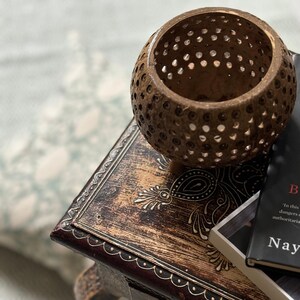 May include: A brown, spherical candle holder with many small holes rests on a decorative wooden box. The box has ornate carvings and a dark finish. A book with a black cover is partially visible.