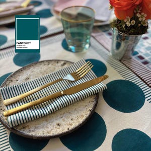 May include: A table setting featuring a plate, gold fork and knife, and a striped napkin. The plate has a speckled design. The tablecloth has a pattern of large teal circles. A glass of water and a small bucket of flowers are also present.