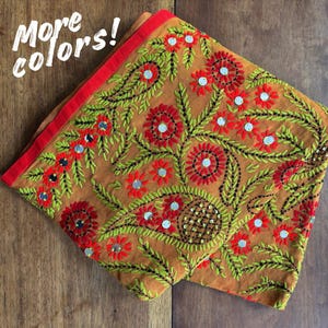 Peut inclure: Textile brodé plié, de couleur beige, avec des motifs floraux rouges et des feuilles vertes. Le motif comprend de petits miroirs réfléchissants. La bordure est rouge et le texte "More colors!" est visible.