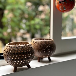 May include: Two brown, spherical candle holders with a textured, perforated design. Each holder has three small wooden legs. The candle holders are displayed on a white windowsill with a blurred green background.