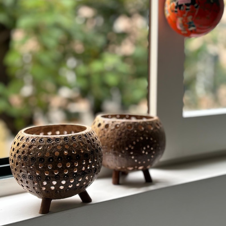 May include: Two brown, spherical candle holders with a textured, perforated design. Each holder has three small wooden legs. The candle holders are displayed on a white windowsill with a blurred green background.