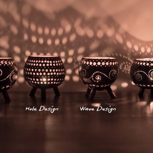 May include: Four black candle holders with detailed patterns and small legs. The candle holders feature a hole design and a wave design. The lit candles cast a pattern of light on the wall behind them.
