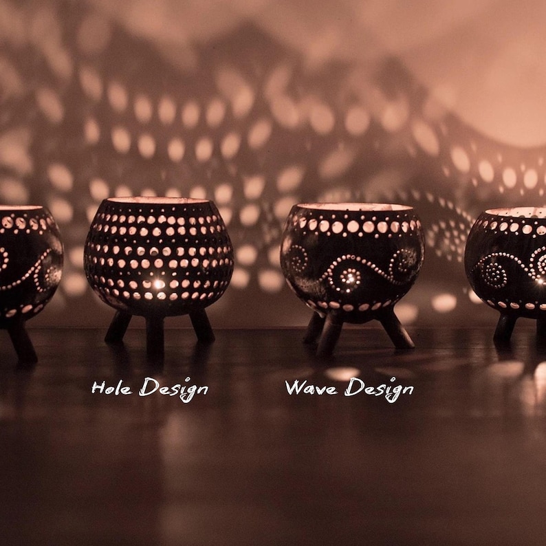 May include: Four black candle holders with detailed patterns and small legs. The candle holders feature a hole design and a wave design. The lit candles cast a pattern of light on the wall behind them.