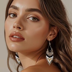 May include: A woman wearing silver-toned dangle earrings. The earrings have a black teardrop shape at the top, with a geometric pattern of white, black, and gold beads. The earrings are a statement piece.