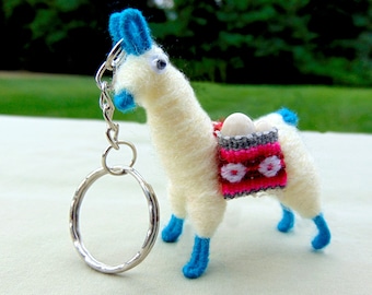 May include: White felt llama keychain with blue ears and hooves. The llama is carrying a small pink and gray bag with white beads.