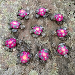 May include: Ten small ceramic sea turtles with pink and green shells. The turtles are arranged in a circle on a brown and green mossy surface.