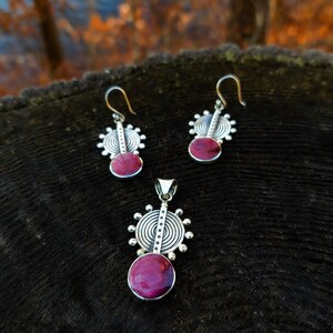 May include: A set of silver earrings and a pendant necklace with a circular design and a pink stone in the center.