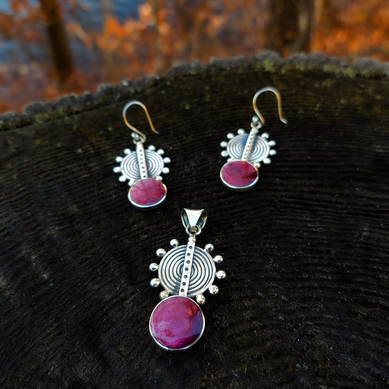 May include: A set of silver earrings and a pendant necklace with a circular design and a pink stone in the center.